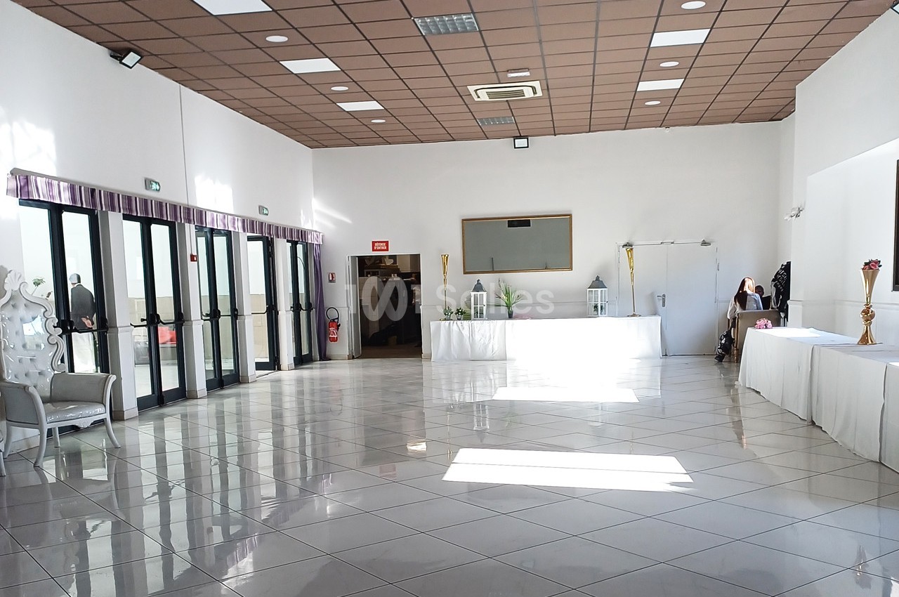 Salle lumineuse avec carrelage blanc, grandes baies vitrées, tables décorées et plafond à dalles marron.