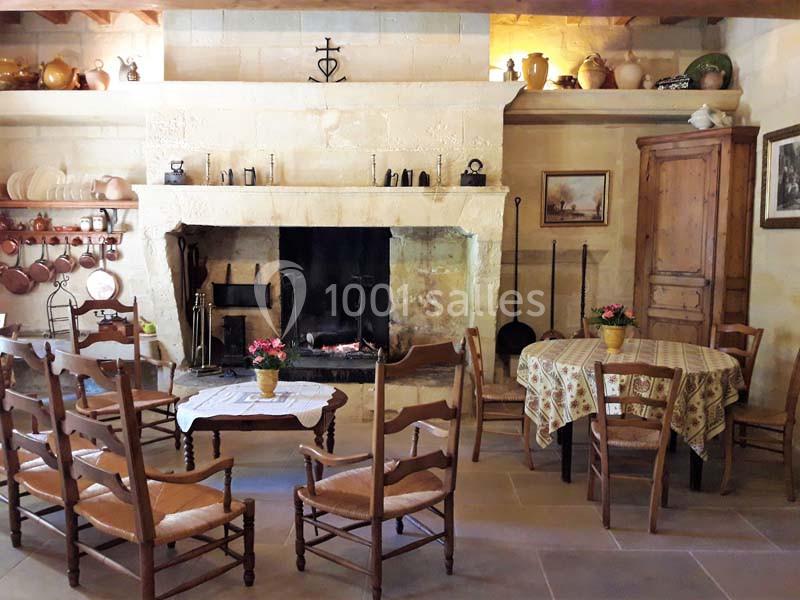 Cuisine rustique avec cheminée en pierre, meubles en bois, table ronde dressée et ustensiles anciens accrochés au mur.