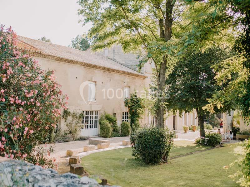 Façade d'une maison en pierre entourée d'un jardin arboré et fleuri sous un ciel dégagé.