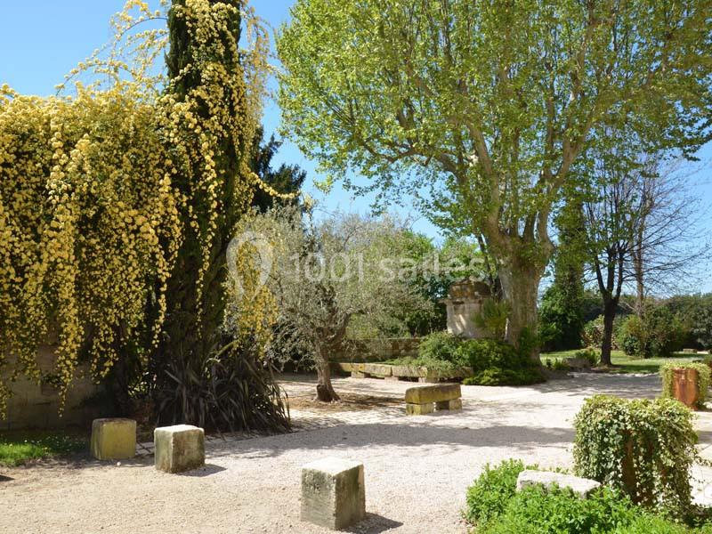 Jardin arboré avec un arbre fleuri jaune, des bancs en pierre et des arbres verdoyants sous un ciel dégagé.
