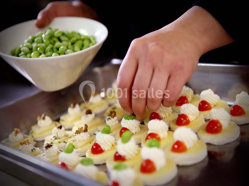 Une main décore des amuse-bouches avec des fèves vertes, des tomates cerises et de la crème sur un plateau en métal.