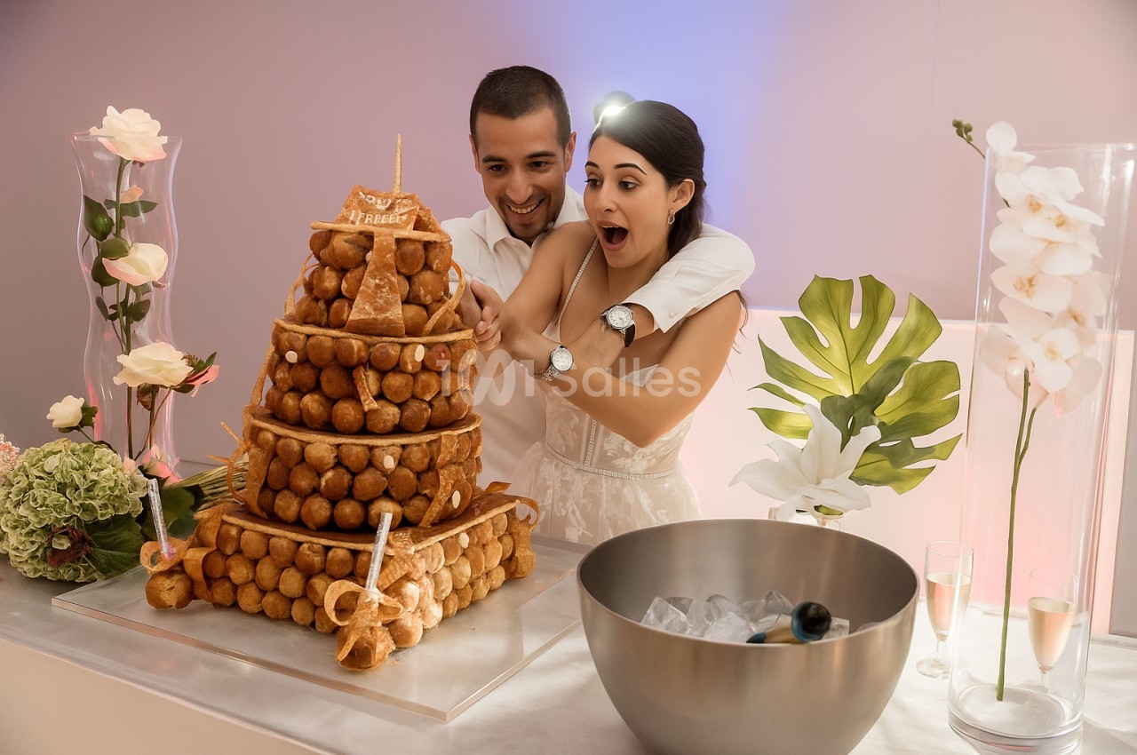 Un couple souriant coupe une pièce montée en choux lors d'une célébration, entouré de décorations florales.