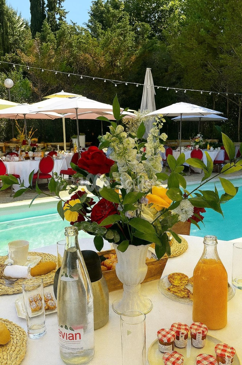 Centre de table fleuri près d'une piscine, entouré de tables dressées avec parasols dans un cadre extérieur verdoyant.