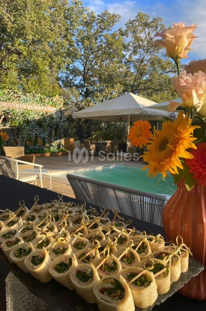 Plateau de bouchées roulées garnies, disposé près d'une piscine entourée de fleurs et de mobilier de jardin.