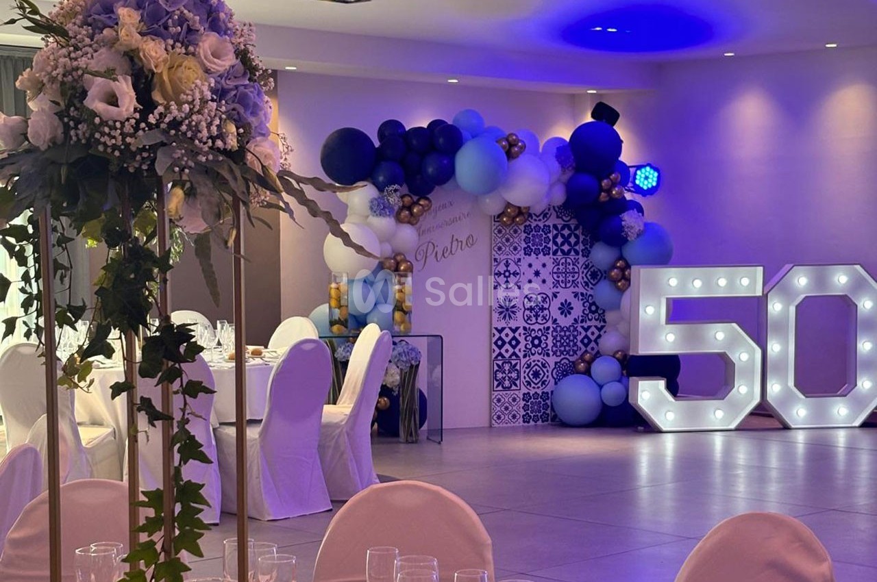Salle décorée pour une fête avec ballons bleus et blancs, chiffre lumineux ’50’ et tables dressées avec fleurs.