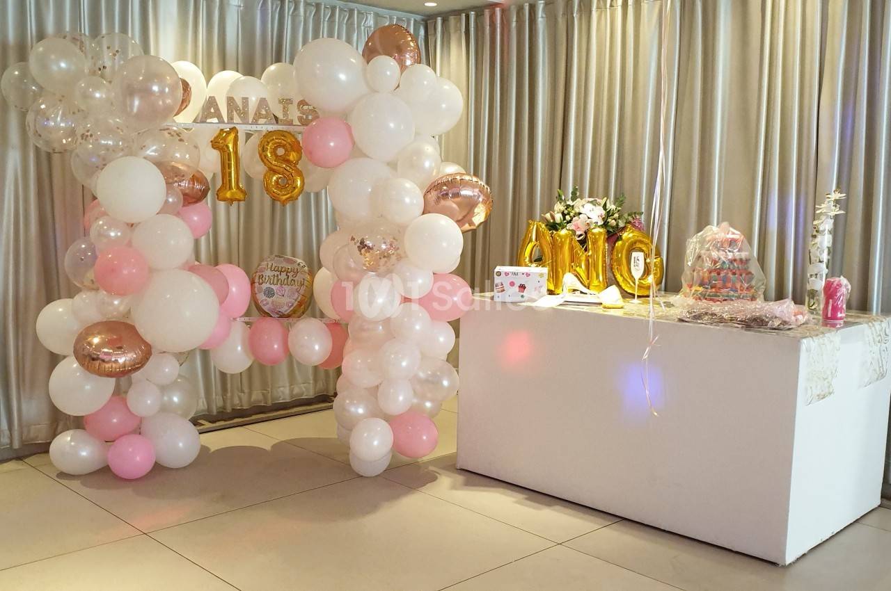 Décoration de fête avec arche de ballons roses et blancs, table ornée de fleurs, gâteaux et éléments festifs.
