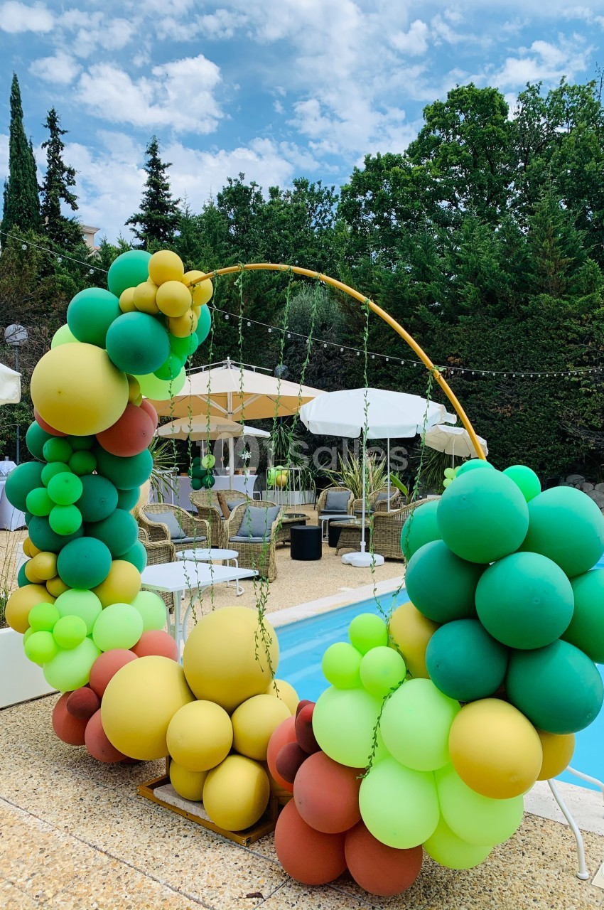 Arc décoratif en ballons colorés près d'une piscine, entouré de chaises et parasols dans un cadre extérieur verdoyant.