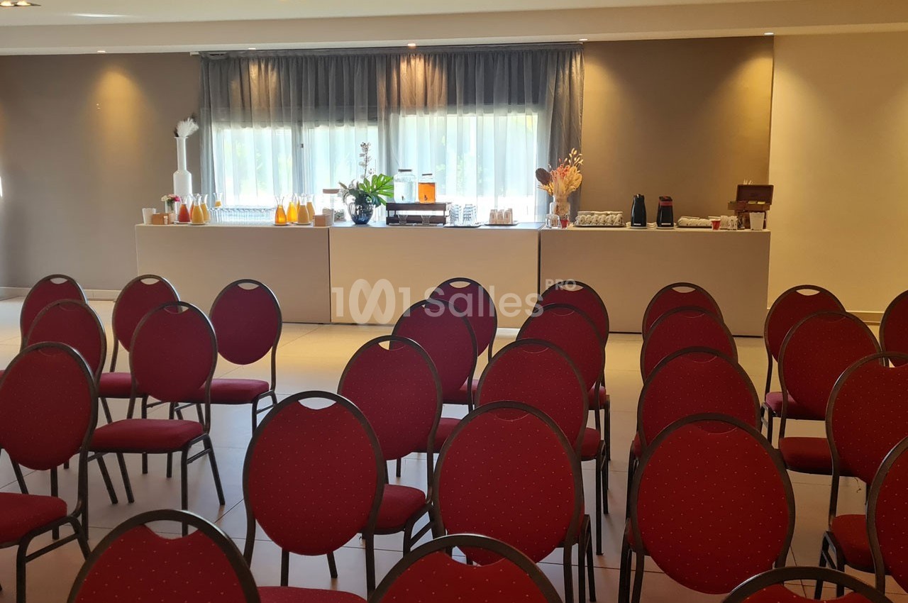 Salle de conférence avec des rangées de chaises rouges et une table au fond proposant des boissons et collations.