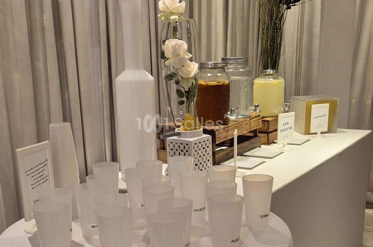 Table avec des boissons en distributeurs, verres en plastique translucides et décorations florales blanches.