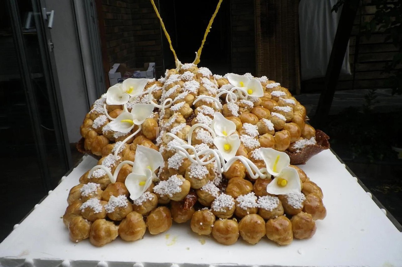 Pièce montée en choux garnis de crème, décorée de fleurs en sucre et de glaçage blanc, posée sur un support blanc.