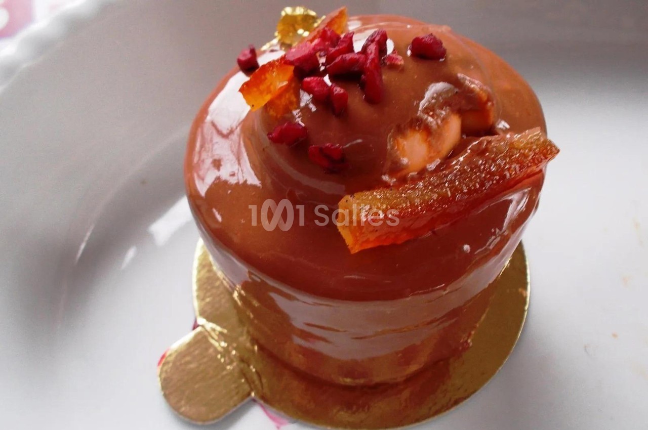 Pâtisserie individuelle glacée au caramel, décorée de morceaux d'orange confite et éclats rouges, sur un support doré.