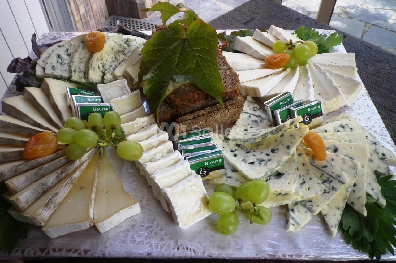 Plateau de fromages variés avec raisins, abricots secs, beurre et pain, présenté sur un plateau décoré.
