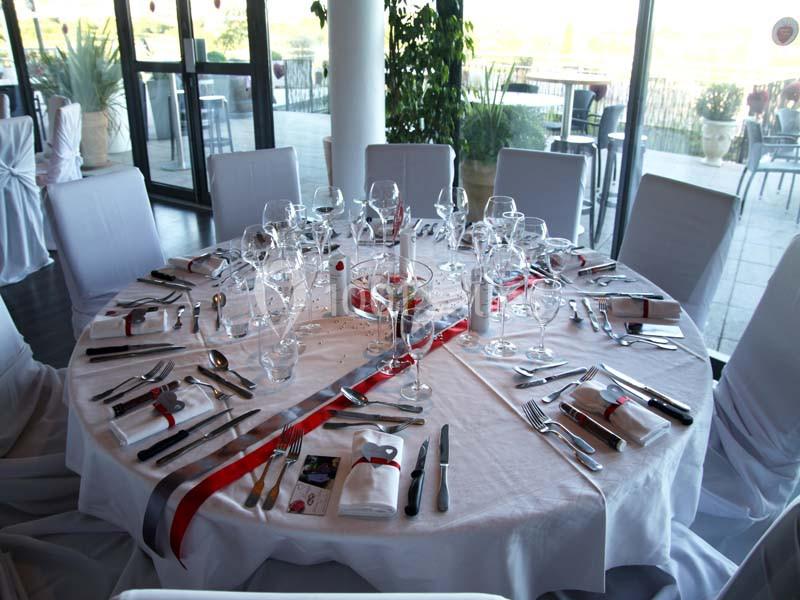 Table ronde dressée en blanc avec couverts, verres et rubans rouges, baies vitrées et terrasse en arrière-plan.