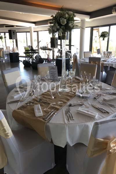 Salle de réception avec tables rondes dressées en blanc et or, compositions florales élevées, baies vitrées et colonnes blanches.