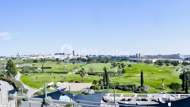 Parcours de golf verdoyant avec fairways et greens, skyline urbain en arrière-plan, vue depuis la terrasse du club-house.