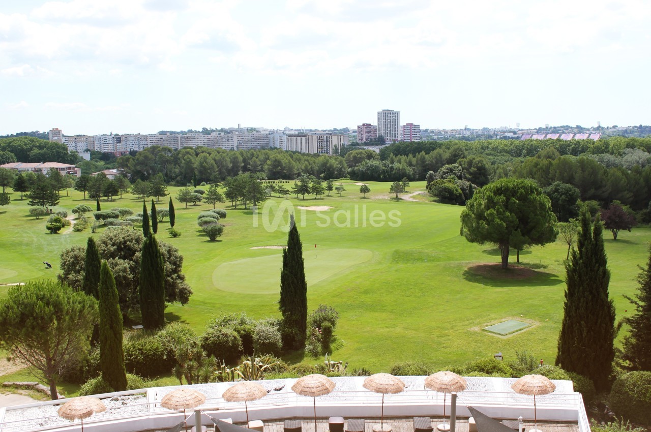 Terrasse ombragée surplombant un parcours de golf avec cyprès, arbres et skyline urbaine.