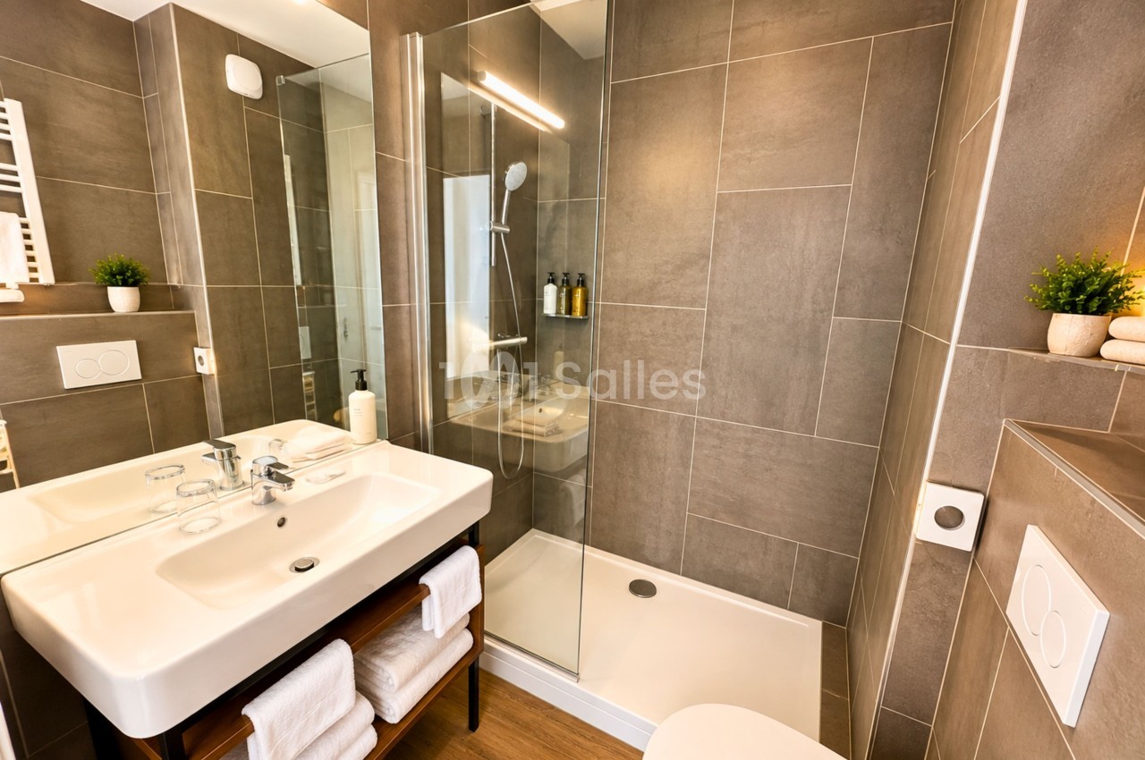 Salle de bains avec carrelage taupe, double vasque blanche, douche à l'italienne vitrée et éclairage LED.