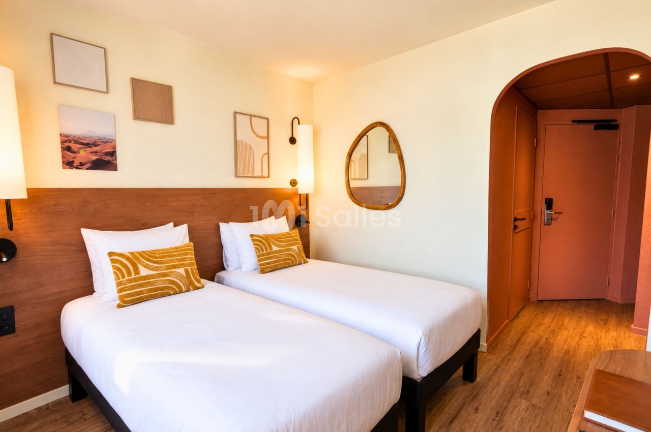 Chambre avec deux lits simples côte à côte, tête de lit en bois, coussins jaunes, miroir ovale, murs beige et porte orange.