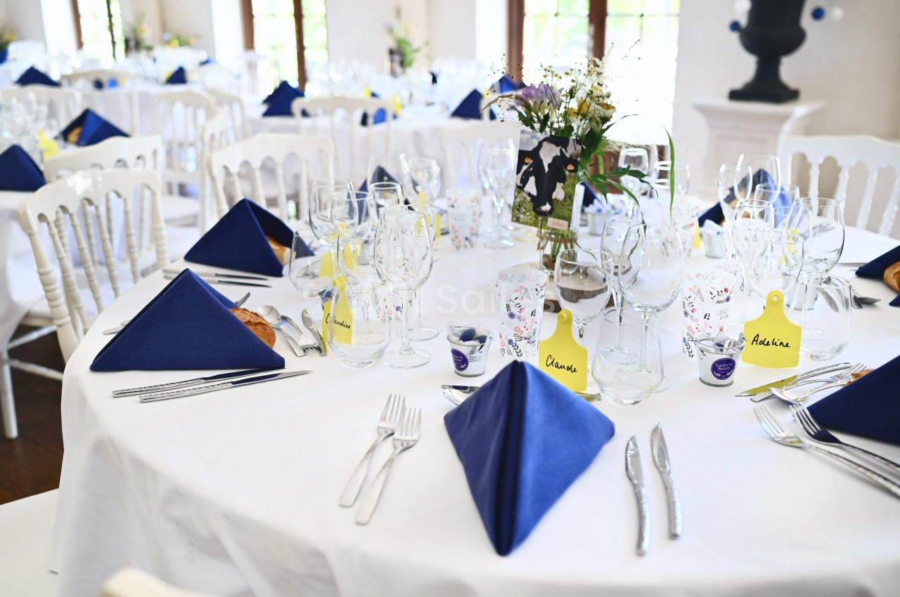 Tables dressées pour un événement avec nappes blanches, serviettes bleues pliées, verres et décorations florales.