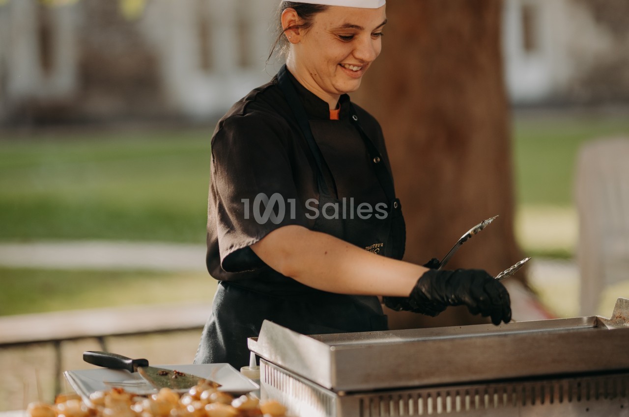 Une cuisinière en tenue noire utilise des pinces pour préparer des aliments sur une plaque chauffante en extérieur.