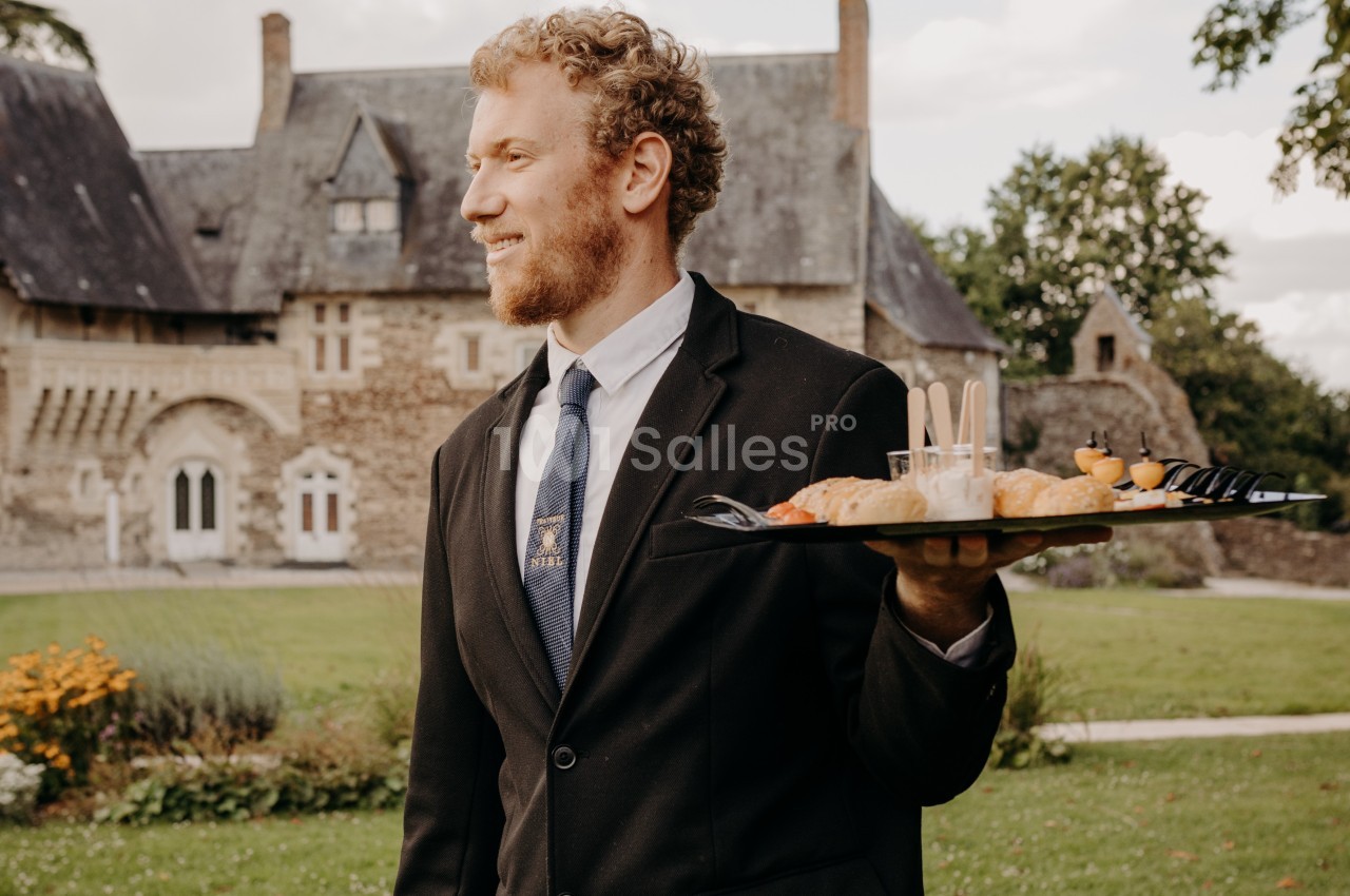Serveur en costume portant un plateau de canapés dans un jardin devant un bâtiment en pierre de style ancien.