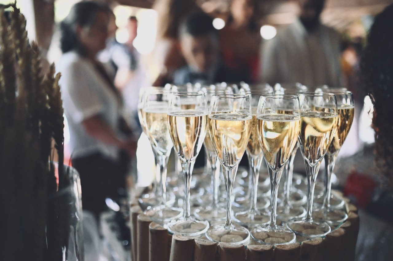 Verres de champagne alignés sur une table en bois lors d'un événement avec des invités en arrière-plan flou.