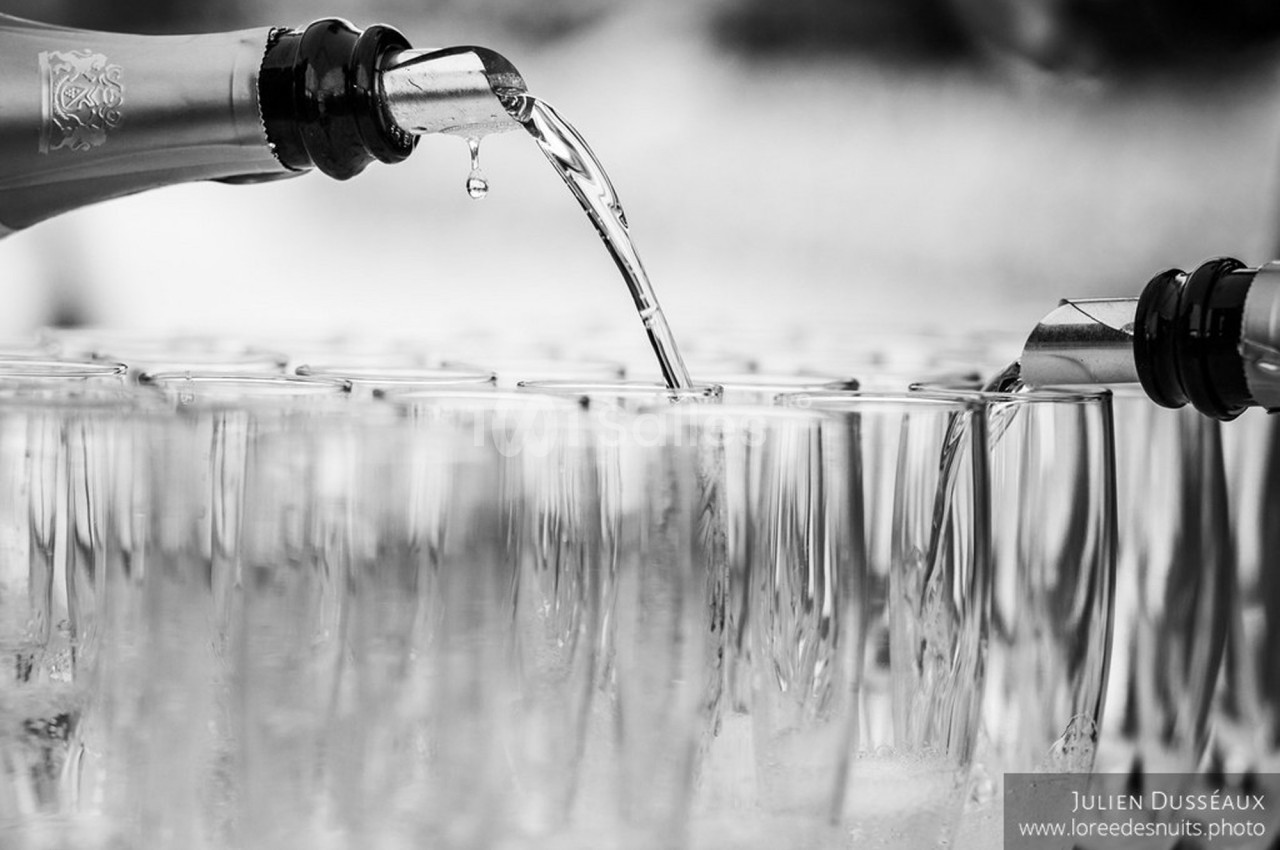 Deux bouteilles versent un liquide transparent dans des rangées de verres alignés, en noir et blanc.