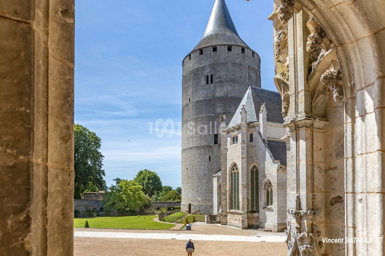 Vue d'une tour médiévale et d'une église entourées de verdure, avec une personne marchant sur une place dégagée.