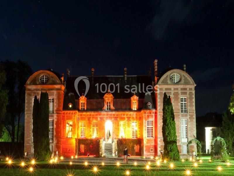 Façade d'un château illuminé en orange dans la nuit, entouré d'arbres et de lumières au sol.