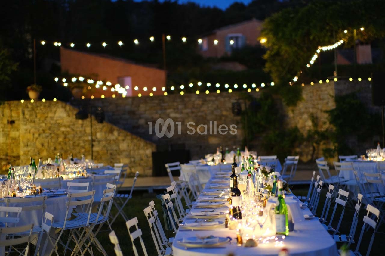 Tables dressées avec nappes blanches et guirlandes lumineuses dans un jardin en soirée.