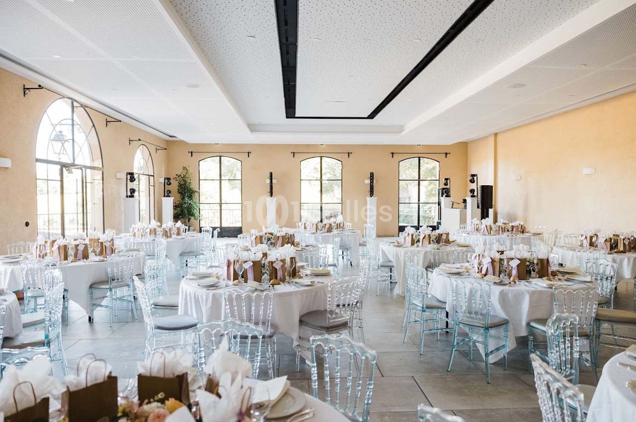 Salle de réception lumineuse avec tables rondes dressées, chaises transparentes et grandes fenêtres en arc.