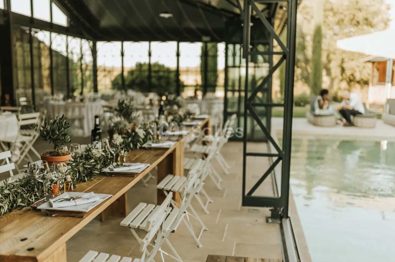 Salle lumineuse avec longues tables en bois décorées de feuillage, donnant sur une piscine et un espace extérieur verdoyant.