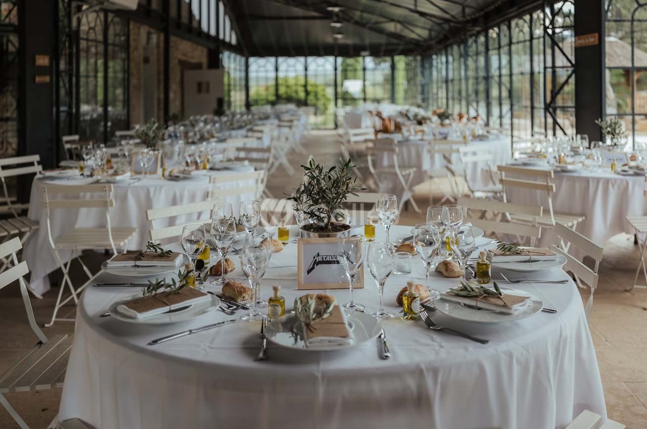Salle lumineuse avec tables rondes dressées pour un repas, décorées de nappes blanches et centres de table végétaux.