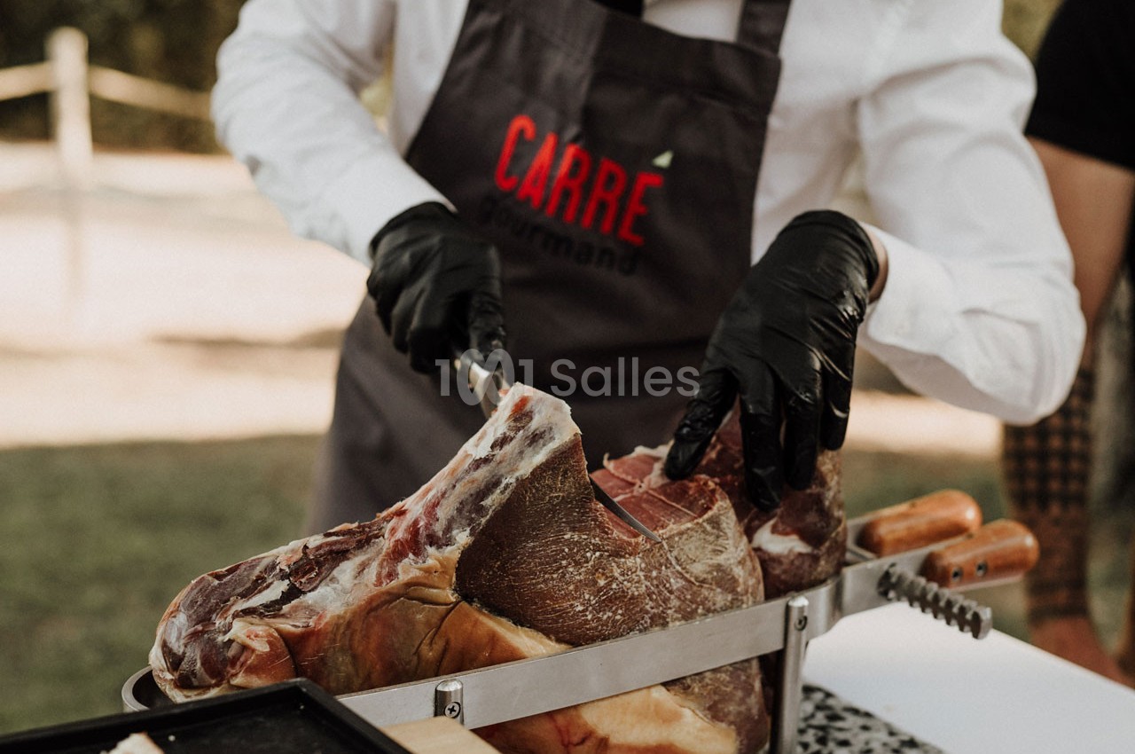 Un cuisinier en tablier découpe un jambon sec à l'aide d'un couteau sur un support en extérieur.