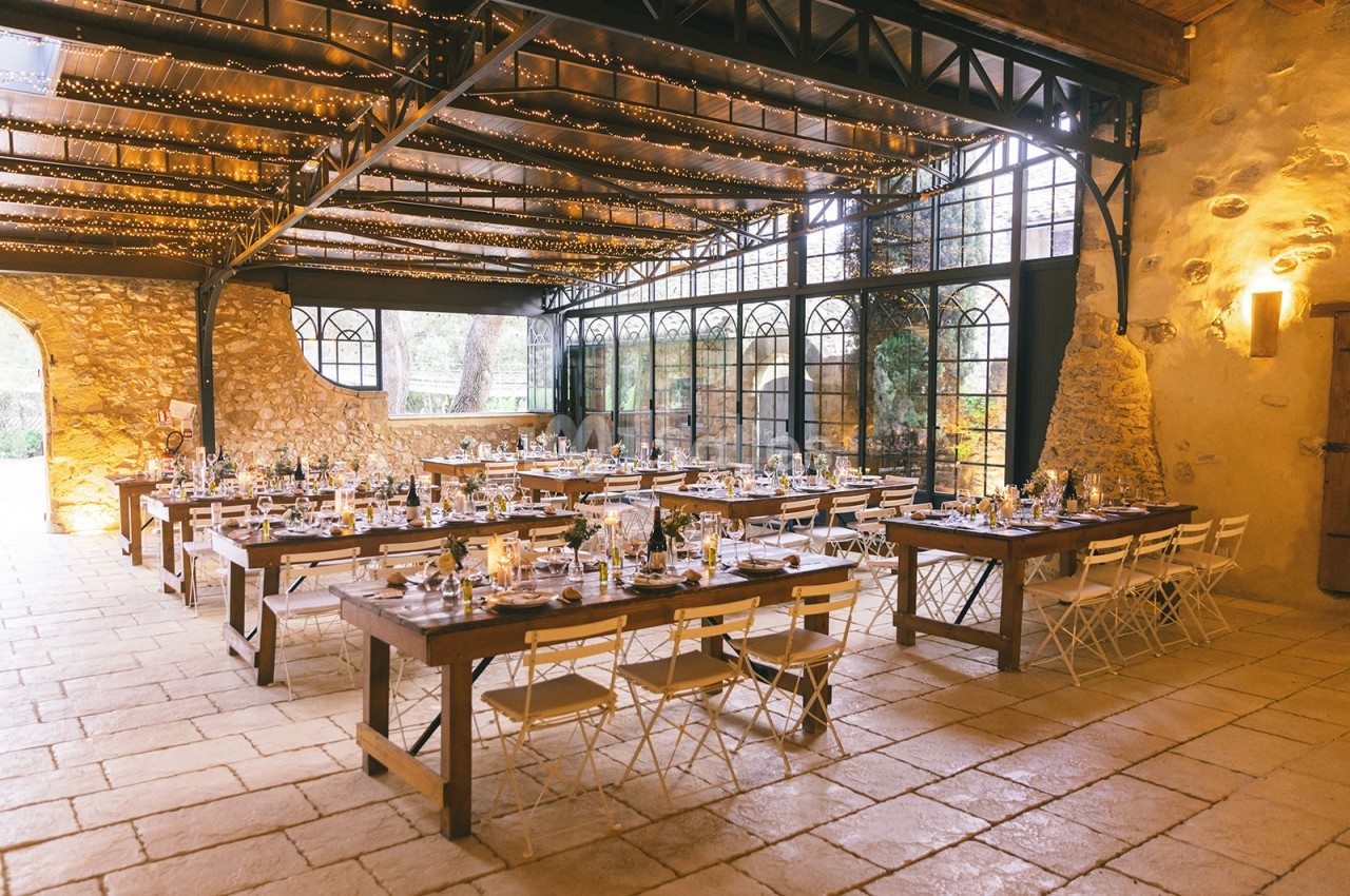 Salle de réception lumineuse avec tables en bois dressées, chaises blanches et murs en pierre apparente.