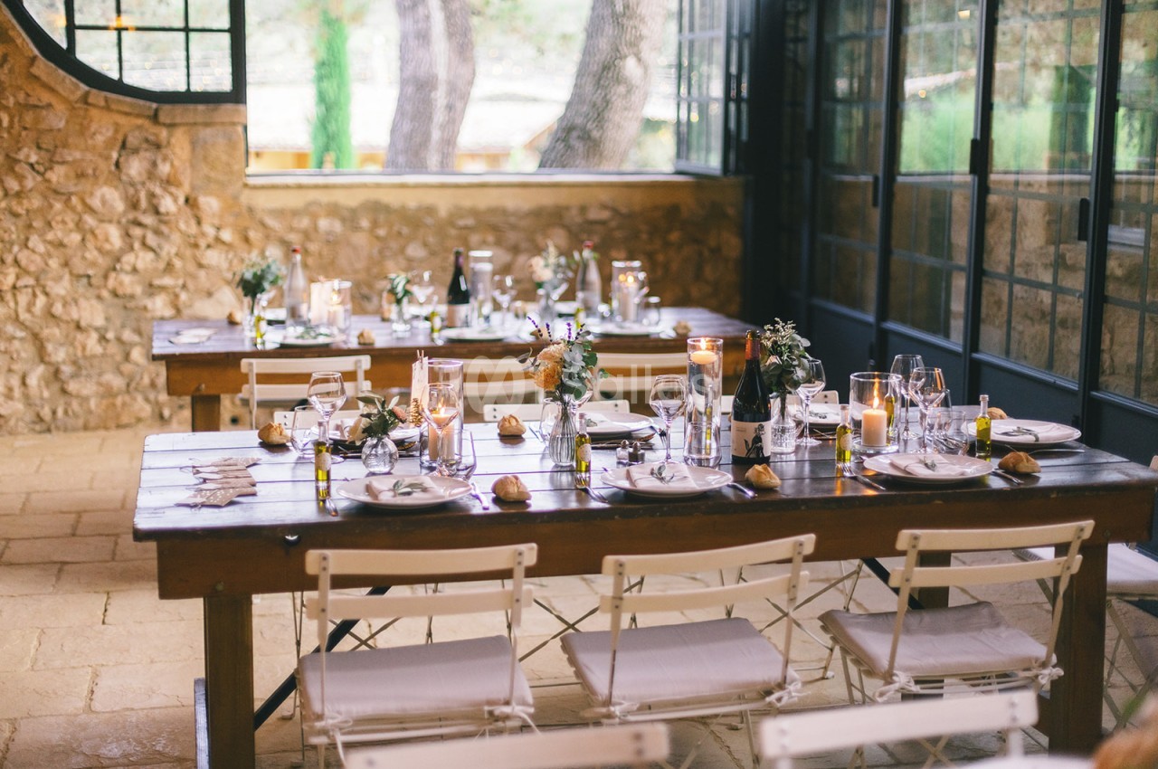 Tables en bois dressées avec vaisselle, bougies et décorations florales dans une salle lumineuse aux murs en pierre.
