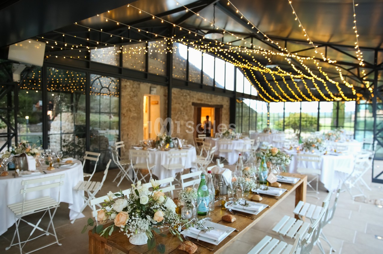 Salle de réception décorée avec des guirlandes lumineuses, des tables dressées et des chaises blanches.
