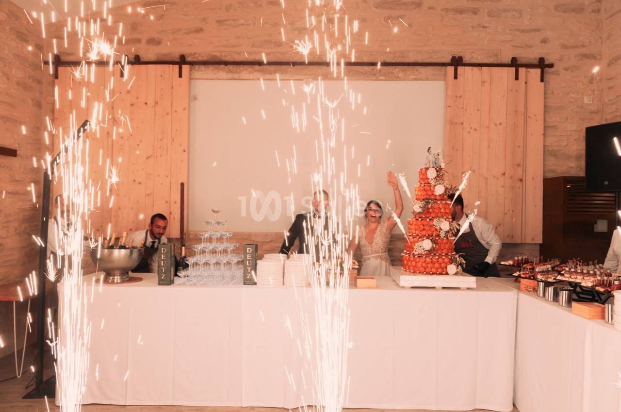 Des étincelles jaillissent devant une table de réception avec une pièce montée, des verres empilés et des convives souriants.