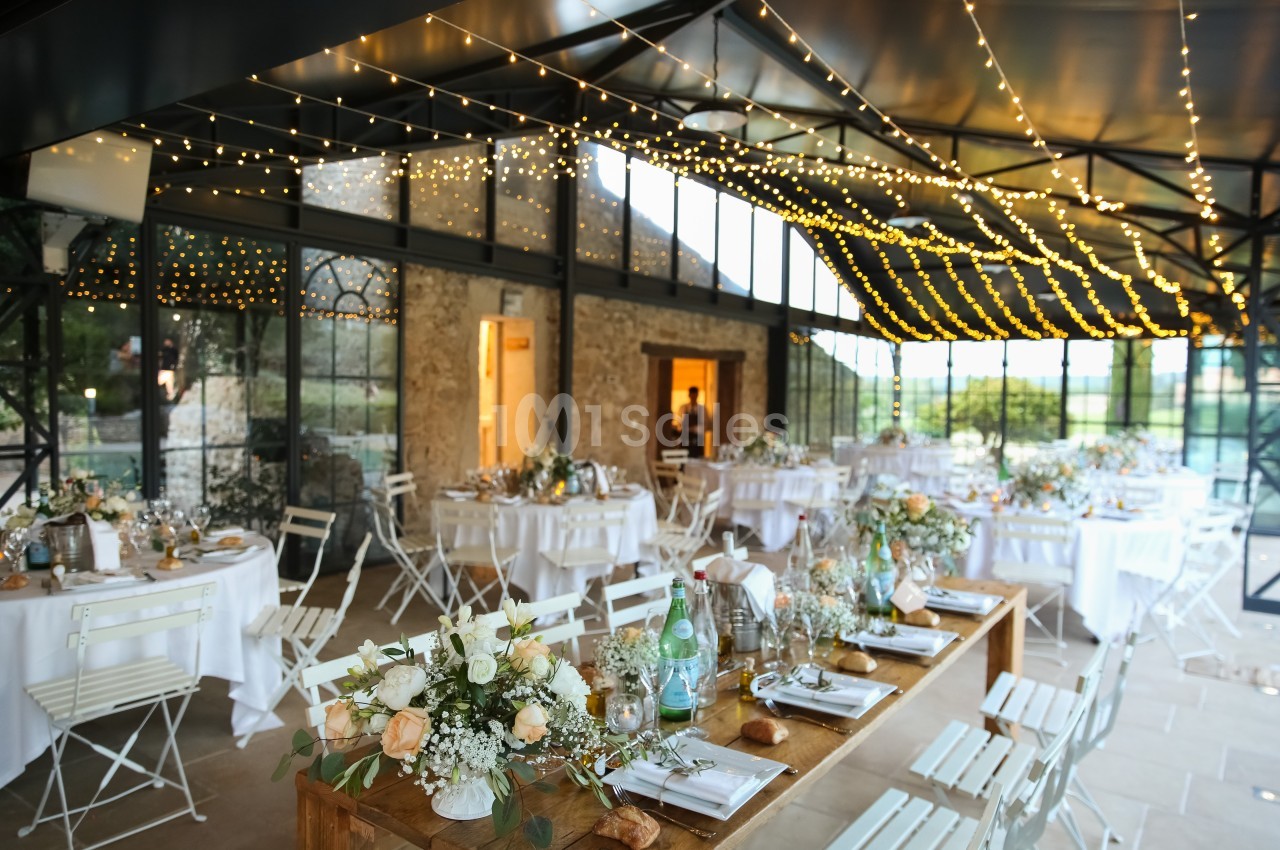 Salle de réception décorée pour un mariage, avec tables dressées, guirlandes lumineuses et grandes baies vitrées.