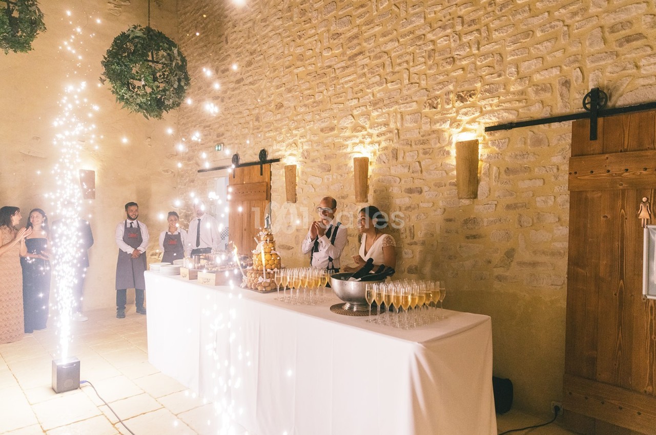Table de réception avec champagne, pièce montée et invités dans une salle en pierre éclairée par des lumières festives.