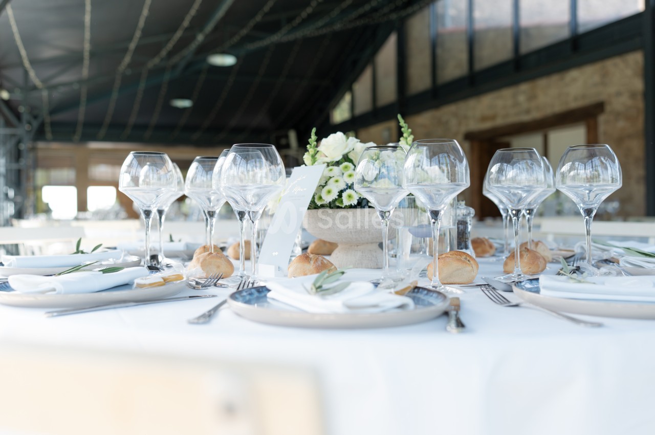 Table dressée avec nappes blanches, verres à vin, couverts, petits pains et centre de table floral dans une salle lumineuse.