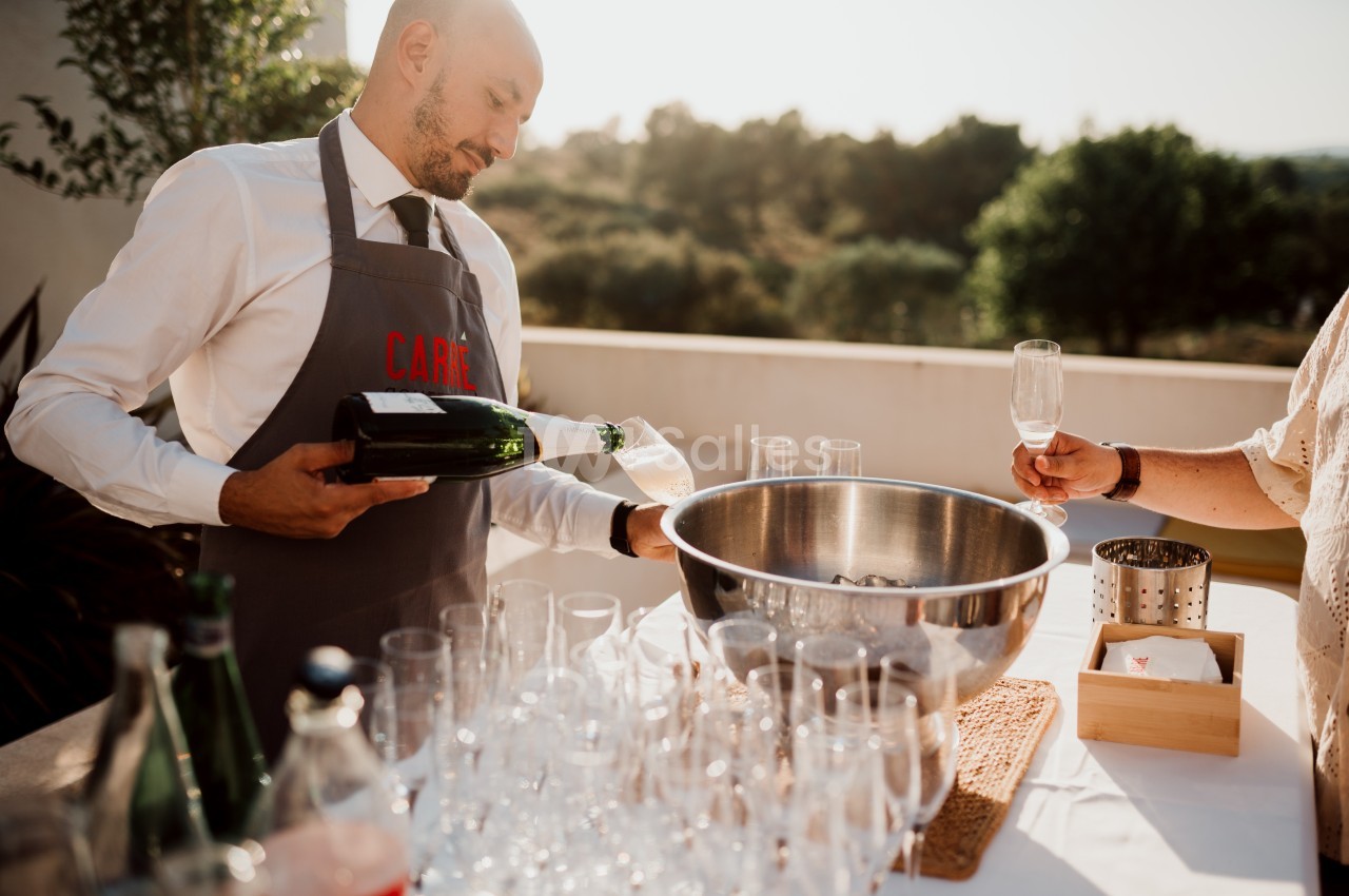Un serveur verse du champagne dans une flûte lors d'un événement en extérieur, avec des verres alignés sur une table.
