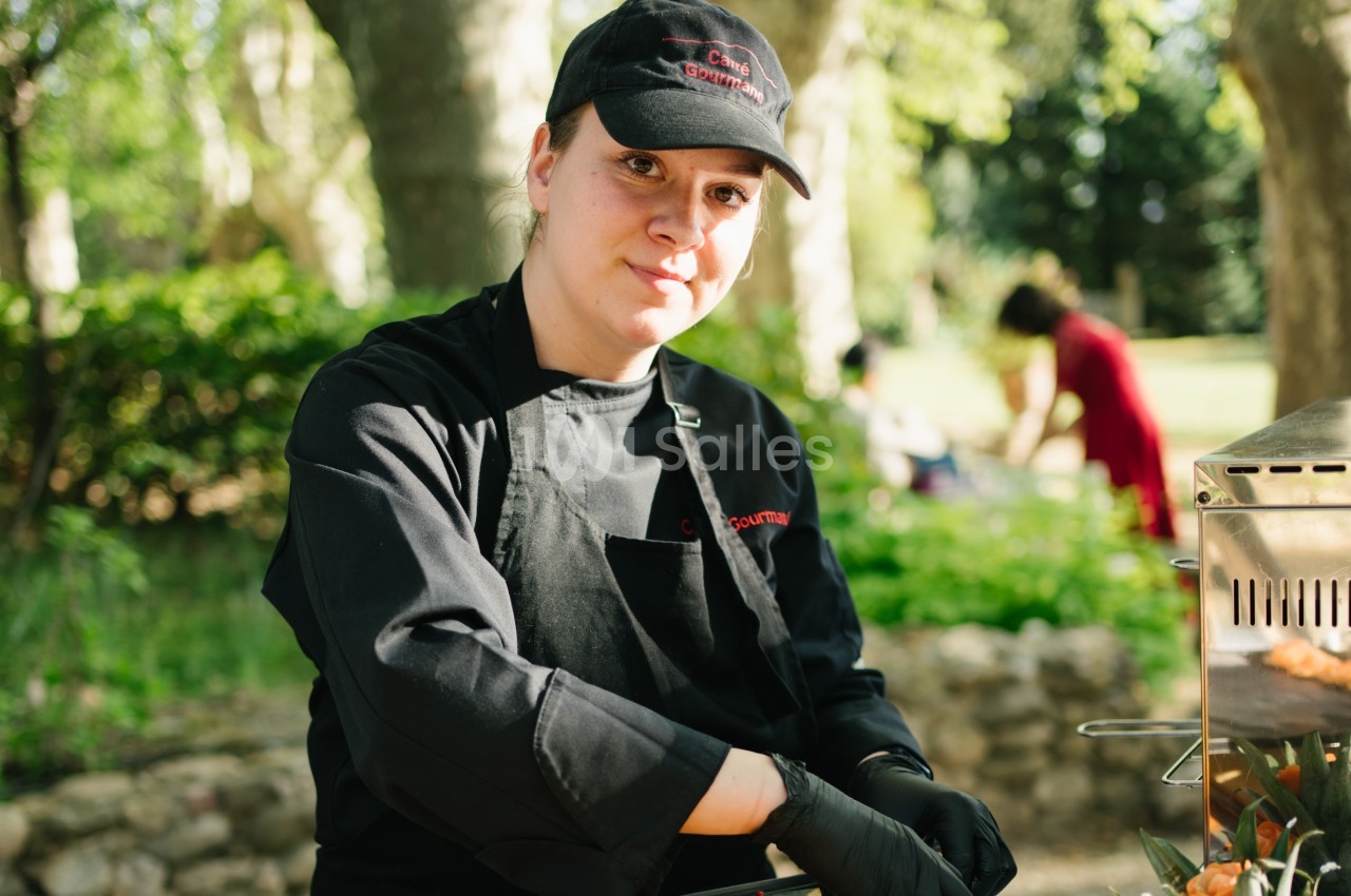 Une cuisinière en tenue noire prépare des aliments en plein air, avec un fond de verdure et de lumière naturelle.
