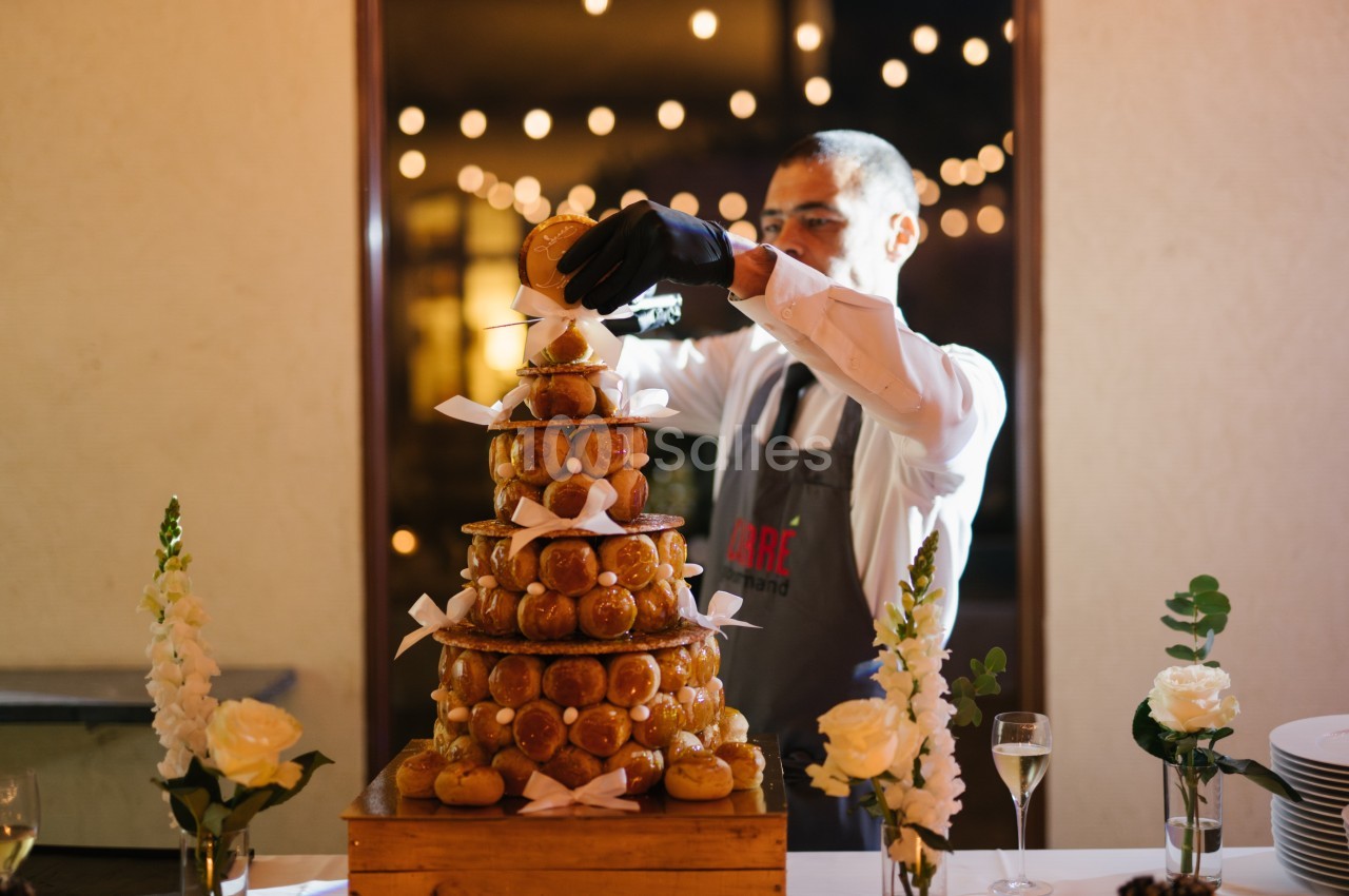 Un serveur assemble une pièce montée de choux décorée de rubans blancs, entourée de fleurs et de lumières.