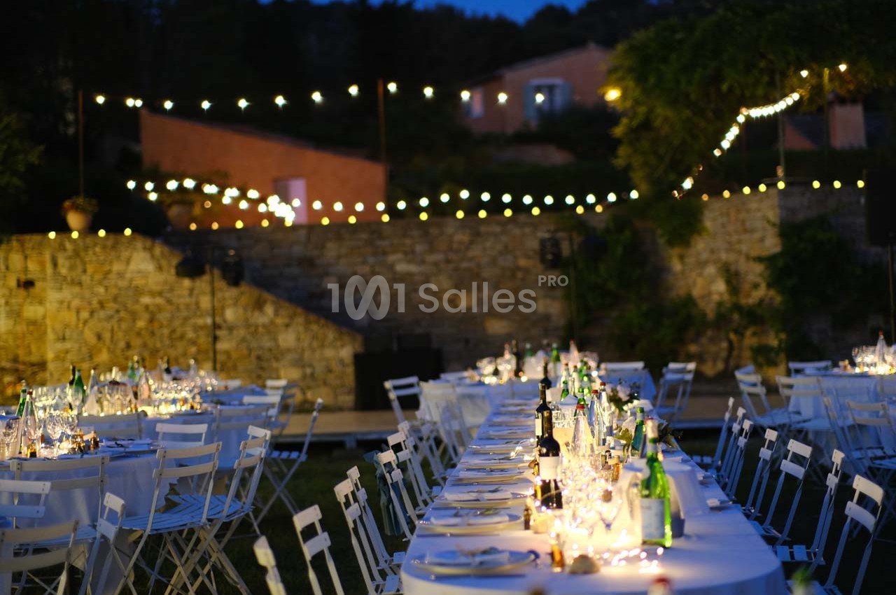 Tables dressées avec nappes blanches et guirlandes lumineuses dans un jardin en soirée.