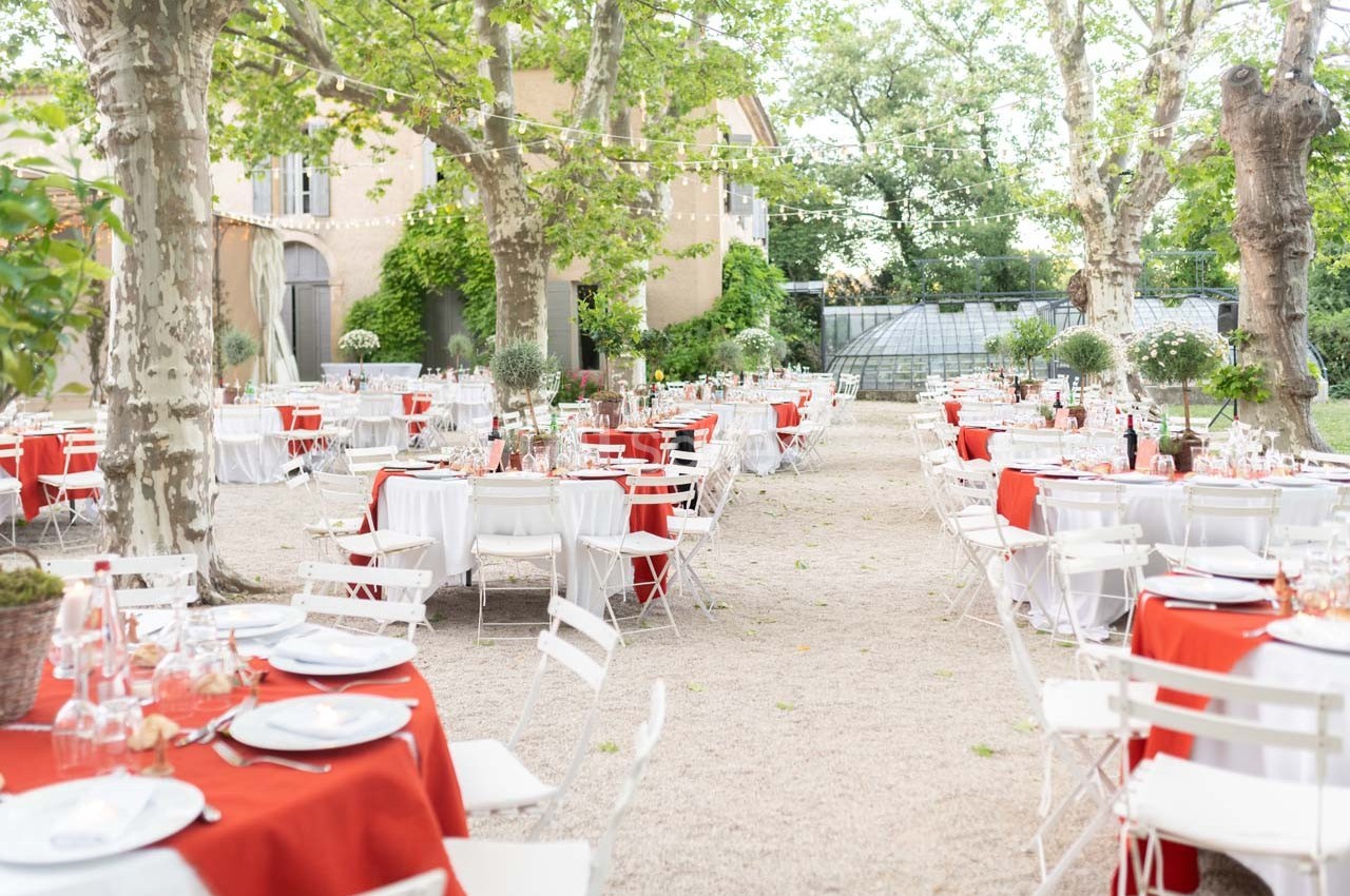 Tables dressées avec nappes rouges et blanches dans un jardin ombragé, entourées de chaises blanches et décorations…