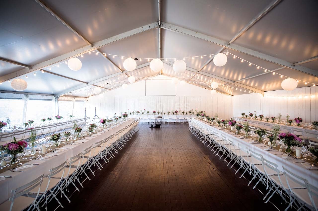 Grande salle de réception sous une tente, avec de longues tables décorées de fleurs et éclairées par des guirlandes…