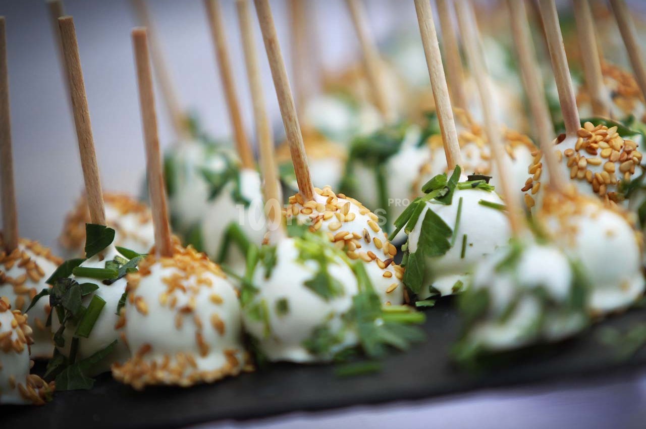 Boules apéritives enrobées de fromage, garnies de graines de sésame et d'herbes fraîches, présentées sur des bâtonnets.