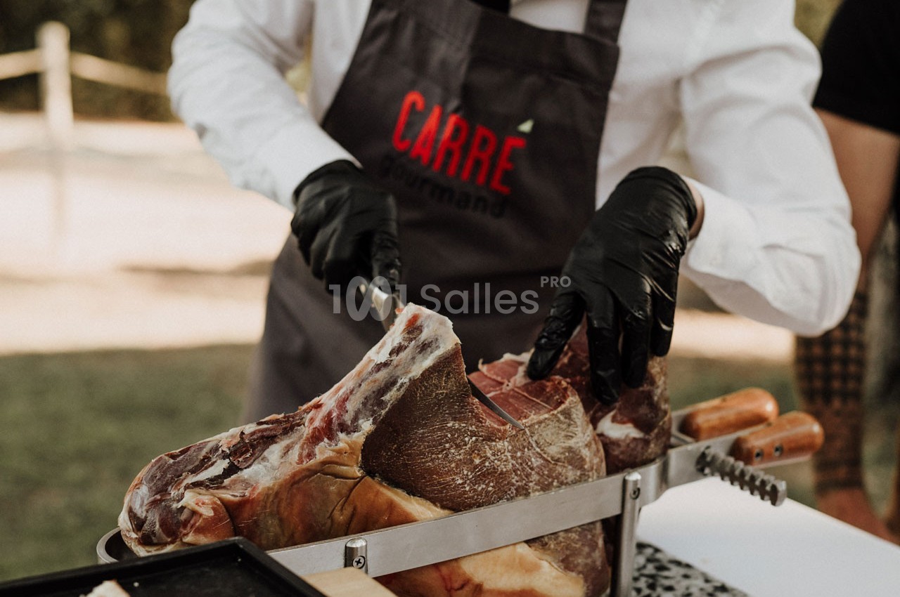 Un cuisinier en tablier découpe un jambon sec à l'aide d'un couteau sur un support en extérieur.