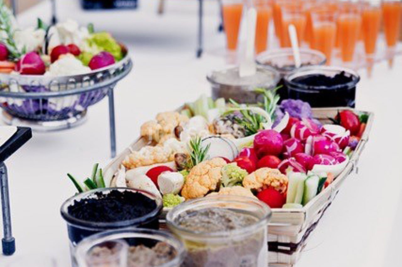 Plateau de légumes frais colorés avec des sauces en pots, entouré de verrines de jus orange sur une table blanche.
